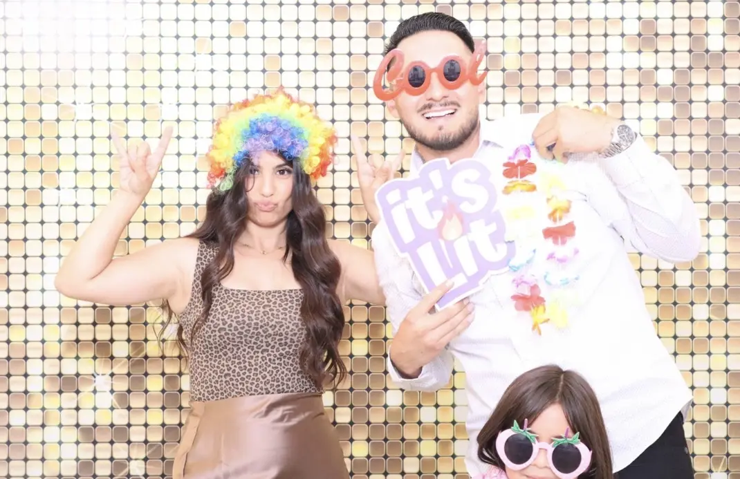 Guests posing with fun props in front of a gold shimmer photo booth backdrop at an event, showing a versatile setup for different celebration styles.