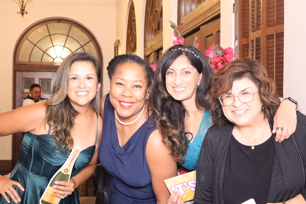 Four smiling women posing warmly together with fun photo booth props, including a champagne bottle cutout, at the Kings and Queens Royale fundraiser at the Ebell Club in Santa Ana.