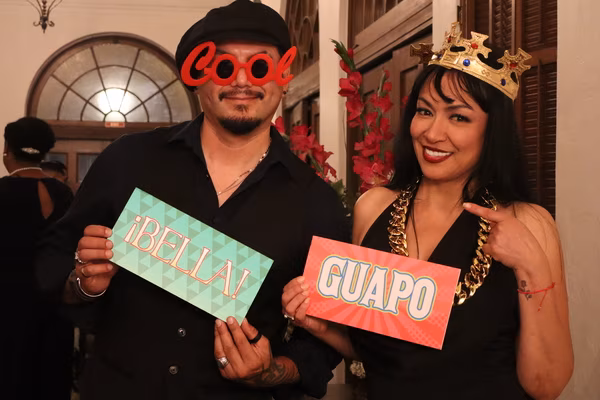 A smiling couple posing with playful photo booth props at the Ebell Club in Santa Ana, with the man wearing red sunglasses holding a "¡BELLA!" sign, and the woman wearing a gold crown holding a "GUAPO" sign.