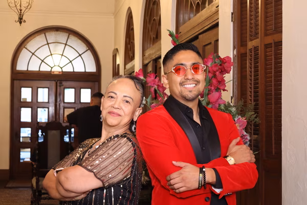 Two elegantly dressed guests posing back-to-back, including a smiling man in a red velvet tuxedo jacket and red sunglasses, standing next to a floral arrangement at the Ebell Club in Santa Ana.