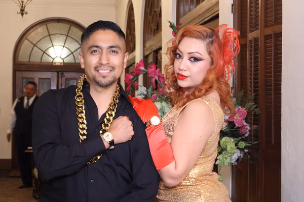 A glamorous couple posing confidently at the Ebell Club in Santa Ana, with the man wearing a black shirt and a thick gold chain prop, and the woman in a gold sequin dress with red gloves.