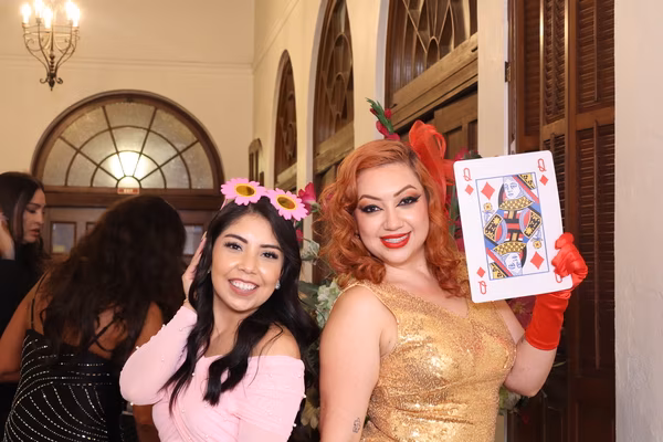 Two smiling female guests at the Ebell Club in Santa Ana, one wearing a pink top and daisy sunglasses, the other in a gold sequin dress and red gloves holding a large Queen of Diamonds playing card prop.