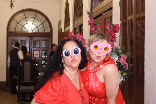 Two women in elegant red dresses making playful kissy faces while wearing fun prop sunglasses—one strawberry-shaped and one pink daisy-shaped—at the Ebell Club in Santa Ana.