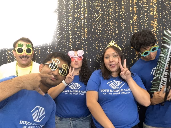 A group of five people, including four wearing blue Boys & Girls Club of the West Valley t-shirts and one wearing a yellow volunteer shirt, posing playfully in front of a black and gold sparkly backdrop. They are wearing fun photo booth props including pineapple sunglasses, oversized dollar sign glasses, pink seashell glasses, a gold tiara, spiky green sunglasses, and holding an inflatable keyboard at Cleveland HS.
