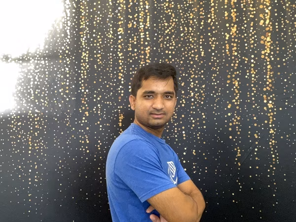 A man wearing a blue Boys & Girls Club t-shirt standing sideways with his arms crossed, looking directly at the camera with a relaxed expression in front of a black backdrop with gold bokeh sparkles at Cleveland HS.