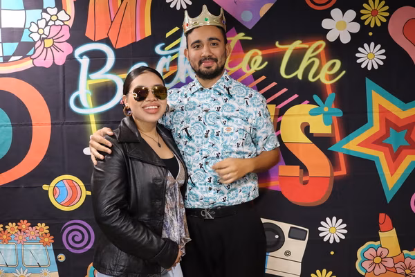 A smiling couple posing together in front of a colorful 70s retro backdrop at Iglesia de Restauración Reseda. The man is wearing a patterned short-sleeve shirt, a black belt, and a gold crown prop, with his arm around the woman, who is wearing a black leather jacket over a patterned top and large tinted sunglasses.