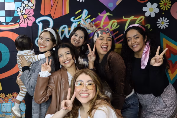 A group of six smiling women and one toddler posing with peace signs in front of a colorful 70s retro backdrop at Iglesia de Restauración Reseda. The women are wearing vintage-inspired outfits, including patterned headbands, a brown leather blazer, and yellow-tinted retro glasses.