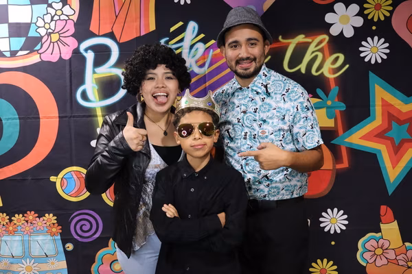A family of three posing in front of a colorful 70s retro backdrop at Iglesia de Restauración Reseda. The woman on the left wears a black curly wig and gives a thumbs up with her tongue playfully sticking out. The man on the right wears a grey fedora and points at the young boy in the center, who stands confidently with his arms crossed, wearing a gold crown and aviator sunglasses.
