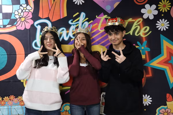 Three smiling young friends posing with peace signs in front of a colorful 70s retro backdrop at Iglesia de Restauración Reseda. They are all wearing crowns, with the two young women in gold tiaras and the young man on the right wearing a large jeweled king's crown.