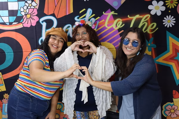 Three smiling women posing in front of a colorful 70s retro backdrop at Iglesia de Restauración Reseda. The women on the left and right are joining arms to form a large heart shape, while the woman in the center makes a smaller heart with her hands. They are wearing vintage-inspired outfits, including a bright rainbow striped shirt, a light-up flower crown, and blue-tinted round sunglasses.