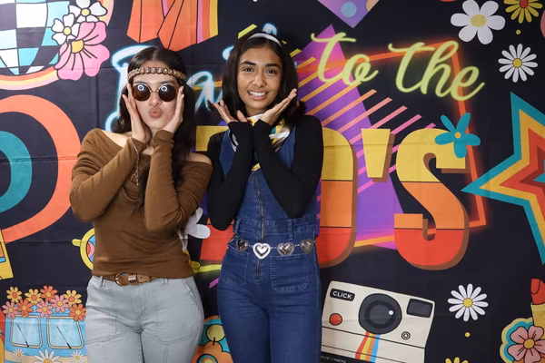 Two young women posing in front of a colorful 70s retro backdrop at Iglesia de Restauración Reseda, framing their faces with their hands. The woman on the left wears a brown off-the-shoulder top, a braided headband, and round retro sunglasses, making a playful kissy face. The woman on the right is smiling broadly and wearing denim overalls with a heart-chain belt and a white headband.