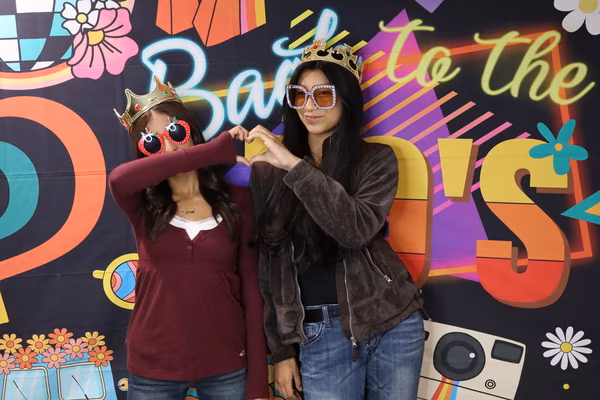 Two young women making a heart shape with their hands together, both wearing gold crowns and fun prop sunglasses, posing in front of a colorful "Back to the 70's" retro backdrop at Iglesia de Restauración Reseda.