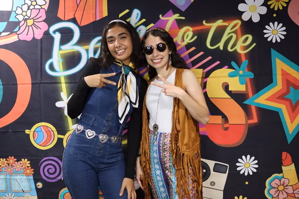 Two smiling women posing with peace signs in front of a colorful 70s retro backdrop at Iglesia de Restauración Reseda. The woman on the left wears denim overalls with a heart-chain belt and a neck scarf, while the woman on the right wears a brown fringed vest, patterned flared pants, and round dark sunglasses.