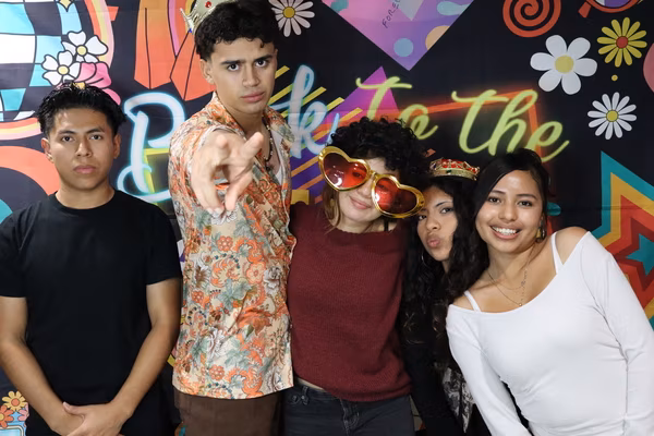 A group of five young people posing in front of a colorful 70s retro backdrop at Iglesia de Restauración Reseda. The two young men on the left are making serious faces, with one wearing a floral shirt and crown while pointing at the camera. The three young women on the right are smiling and making a kissy face, wearing fun props including oversized red heart sunglasses and a gold tiara.