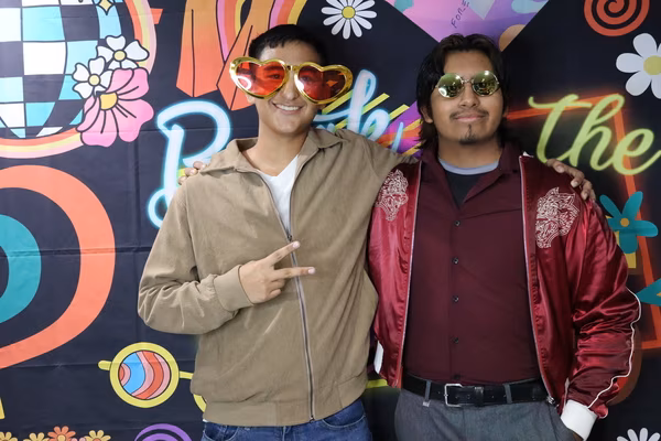 Two smiling young men standing shoulder-to-shoulder in front of a colorful 70s retro backdrop at Iglesia de Restauración Reseda. The man on the left is throwing a peace sign and wearing oversized red heart sunglasses and a tan jacket, while the man on the right wears round vintage sunglasses and a red satin bomber jacket.