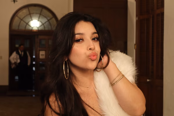 A glamorous female guest with dark hair, large hoop earrings, and a white fur wrap posing playfully with a kissy face near the wooden doors of the Ebell Club in Santa Ana.