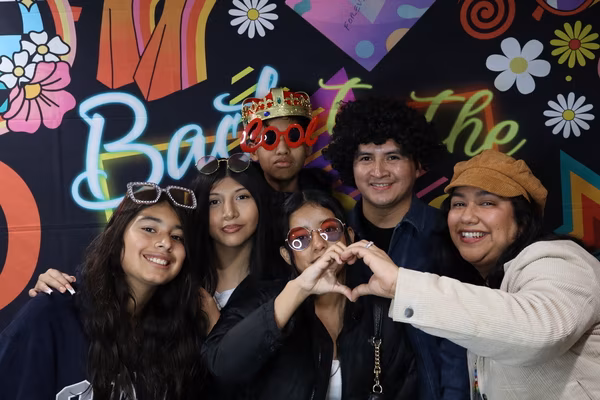 A smiling group of six people posing together in front of a colorful 70s retro backdrop at Iglesia de Restauración Reseda. Two women in the front are making a heart shape with their hands, while others in the group wear fun props including a gold crown, red "COOL" glasses, a curly wig, and vintage sunglasses.