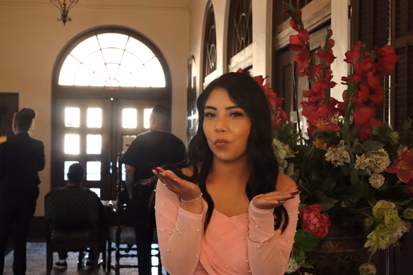 A woman in a pink top with pearl details posing playfully with her hands raised near a large red floral arrangement and arched wooden doors at the Ebell Club in Santa Ana.