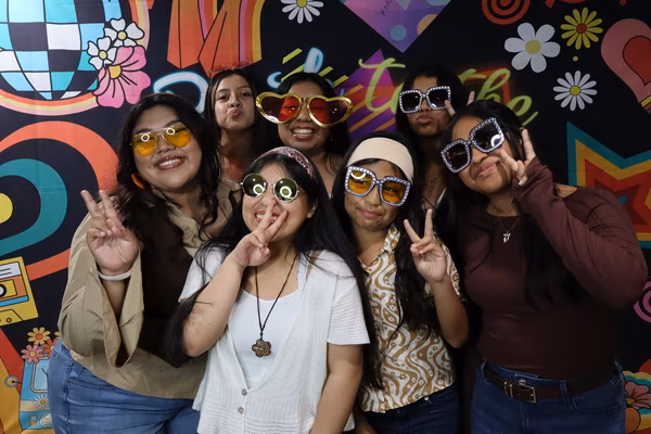 Seven young women smiling and posing with peace signs while wearing various fun prop sunglasses, including oversized hearts and studded frames, in front of a colorful 70s-themed backdrop at Iglesia de Restauración Reseda.