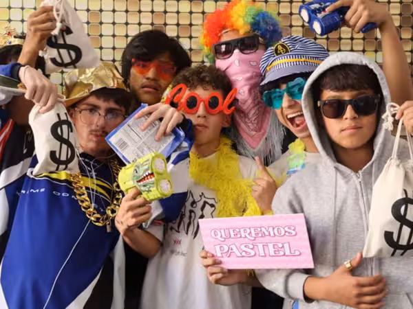 A large, energetic group of teenage boys packed closely together in front of a sparkling gold sequin backdrop at Cleveland HS. They are posing with a chaotic and fun mix of props, including canvas money bags, toy dinosaur guns, a gold chain, a gold hat, a rainbow clown wig with a pink bandana over the face, a sailor hat, red "cool" glasses, and a pink sign that reads "QUEREMOS PASTEL."