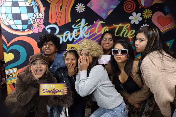A large group of seven smiling friends posing together in front of a colorful 70s retro backdrop at Iglesia de Restauración Reseda. They are wearing fun props, including a curly blonde wig, a curly black wig, and studded square sunglasses, while the woman in the front left playfully holds a sign that says "Saved & Sanctified."