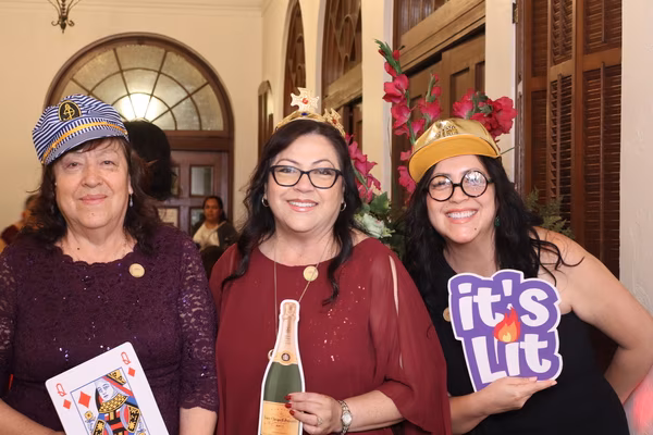 Three smiling women posing warmly together at the Ebell Club in Santa Ana, wearing a sailor hat, a gold crown, and a gold cap, while holding a large Queen of Diamonds card, a champagne bottle cutout, and an "it's Lit" sign prop.
