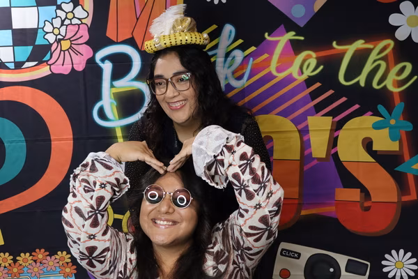 Two smiling friends posing playfully in front of a colorful 70s retro backdrop at Iglesia de Restauración Reseda, with the person in back making a heart shape with her hands over her friend's head. They are wearing fun retro props, including a mini yellow hat and round pink sunglasses.