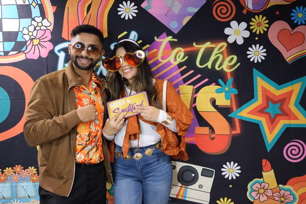 A smiling couple posing in front of a colorful 70s retro backdrop at Iglesia de Restauración Reseda. The man wears a vintage floral shirt, brown jacket, and sunglasses, while the woman wears heart-shaped sunglasses, an orange shawl, and holds a sign reading "Saved & Sanctified."