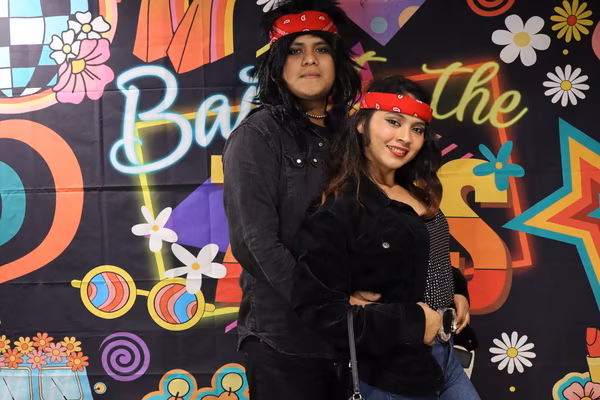 A couple posing together in front of a colorful 70s retro backdrop at Iglesia de Restauración Reseda. They are wearing matching red bandanas. The man is wearing a long black rocker wig and a black shirt, standing behind the smiling woman, who is wearing a black jacket and a sparkly top.