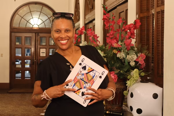 A smiling woman in a black dress making a peace sign while holding a large Queen of Spades playing card prop, standing next to a red floral arrangement and a large inflatable die at the Ebell Club in Santa Ana.