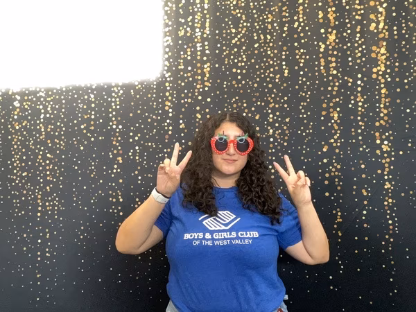 A woman with long curly hair wearing a blue Boys & Girls Club of the West Valley t-shirt and red strawberry-shaped sunglasses. She is smiling gently and holding up two peace signs in front of a black backdrop with gold bokeh sparkles at Cleveland HS.