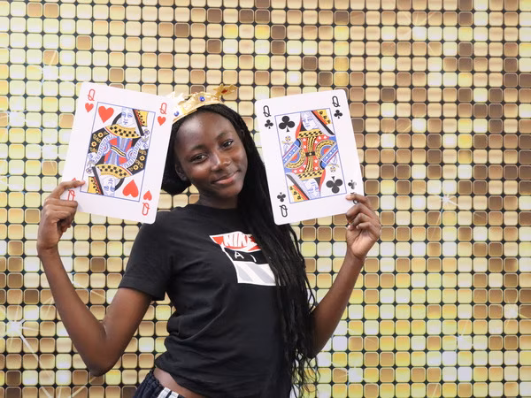 A young woman wearing a pink Polo shirt, dark sunglasses, and a gold crown, holding an oversized Queen of Clubs playing card prop in front of a sparkling gold sequin backdrop at Cleveland HS.