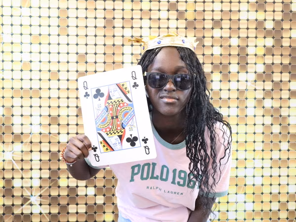 A young woman wearing a pink Polo shirt, dark sunglasses, and a gold crown, holding an oversized Queen of Clubs playing card prop in front of a sparkling gold sequin backdrop at Cleveland HS.