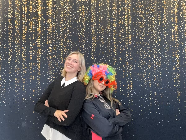 Two young women standing back-to-back with their arms crossed in front of a black backdrop with gold bokeh sparkles at Cleveland HS. The woman on the left is smiling warmly and wearing a black sweater over a white collared shirt. The woman on the right is wearing a black hoodie, a brightly colored rainbow clown wig, and oversized red glasses that spell the word "COOL".