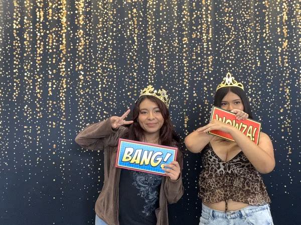 Two young women posing in front of a black backdrop with gold bokeh sparkles at Cleveland HS. The young woman on the left wears a brown jacket and a gold crown, throwing a peace sign while holding a blue comic-style "BANG!" sign. The young woman on the right wears a leopard print top and a gold tiara, playfully hiding her face behind a red comic-style "WOWZA!" sign.