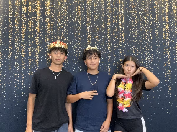 Three students posing in front of a black backdrop with gold bokeh sparkles at Cleveland HS. The young man on the left wears a black t-shirt, a silver cross necklace, and a red and gold crown. The young man in the middle wears a dark blue t-shirt, a silver chain, and a small gold crown while throwing a subtle peace sign. The young woman on the right wears a black t-shirt and a colorful flower lei, playfully framing her face with both hands.