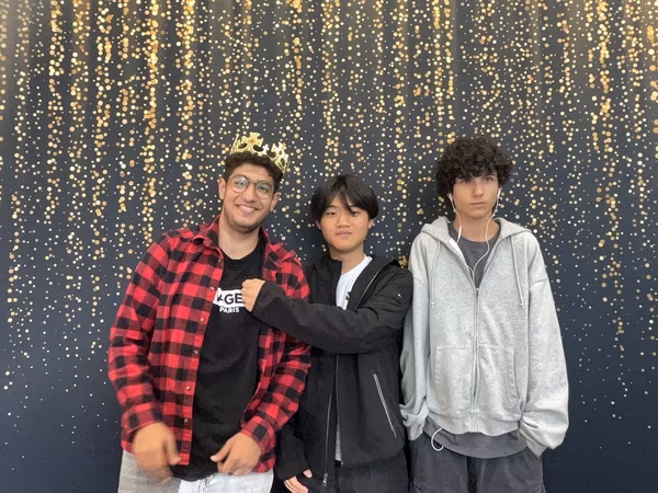 Three young men posing together in front of a black backdrop with gold bokeh sparkles at Cleveland HS. The young man on the left is smiling broadly while wearing a red and black flannel, glasses, and a small gold crown. The young man in the middle is wearing a black jacket and playfully resting his fist on his friend's chest. The young man on the right has a serious expression and is wearing a light grey zip-up hoodie with wired earphones.