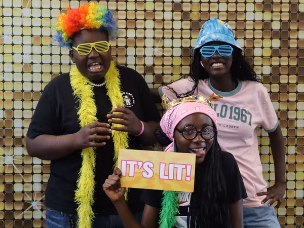 Three students posing playfully in front of a sparkling gold sequin backdrop at Cleveland HS. The student on the left wears a rainbow wig, yellow shutter shades, and a yellow feather boa. The student on the right wears a blue bucket hat and blue shutter shades. The student in the front center wears a pink bandana, a gold crown, round glasses, and a green feather boa while holding a yellow "IT'S LIT!" prop sign.
