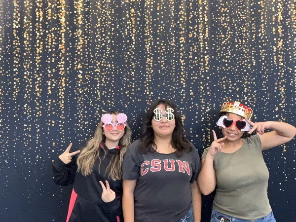 Three young women posing happily in front of a black backdrop with gold bokeh sparkles at Cleveland HS. The young woman on the left wears a black and pink hoodie and pink seashell sunglasses while throwing double peace signs. The young woman in the middle wears a grey CSUN t-shirt and silver dollar sign glasses. The young woman on the right wears an olive green shirt, a red and gold crown, and heart-shaped sunglasses with cartoon hands, while also making peace signs.