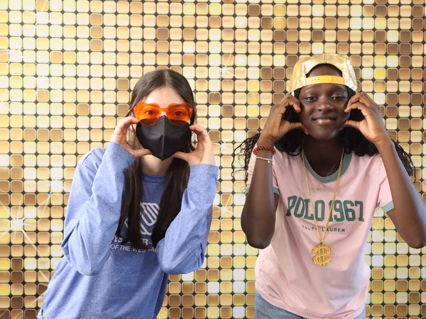 Two young women posing playfully in front of a sparkling gold sequin backdrop at Cleveland HS. The student on the left wears a blue Boys & Girls Club long-sleeve shirt, a black face mask, and orange heart-shaped glasses. The student on the right wears a pink Polo shirt, a backwards gold baseball cap, and a large gold dollar sign necklace, framing her face with her hands.