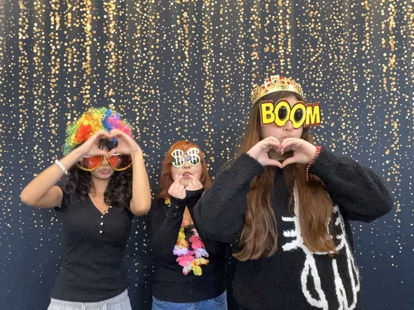 Three young women posing in front of a black backdrop with gold bokeh sparkles at Cleveland HS. The student on the left is wearing a rainbow afro wig and oversized heart sunglasses while making a large heart shape with her hands. The student in the middle is wearing silver dollar sign glasses and a bright flower lei, making a small finger heart. The student on the right is wearing a gold crown and yellow comic-style "BOOM" glasses, also making a heart shape with her hands.
