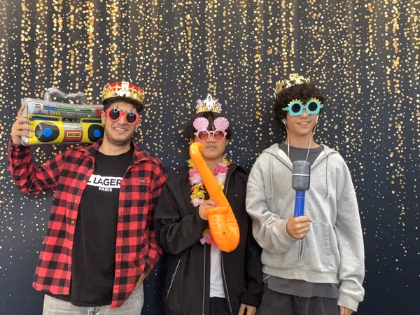 Three young men posing as a musical group in front of a black backdrop with gold bokeh sparkles at Cleveland HS. The young man on the left wears a red flannel, a red and gold crown, and strawberry sunglasses while holding an inflatable boombox on his shoulder. The young man in the middle wears a black jacket, a small crown, pink seashell sunglasses, and a flower lei while holding an orange inflatable saxophone. The young man on the right wears a light grey hoodie, a gold crown, and blue sunflower sunglasses while holding an inflatable microphone.