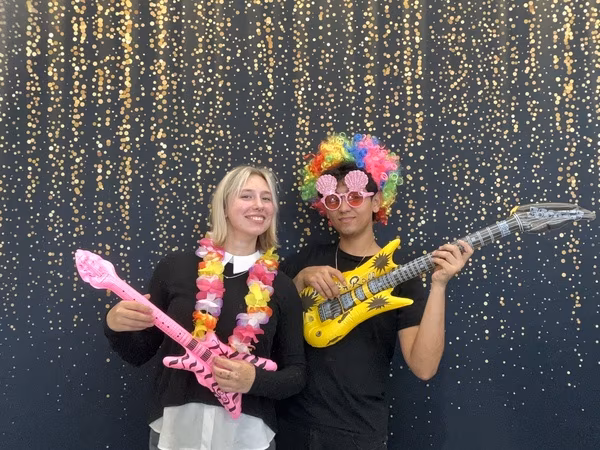 Two students posing playfully in front of a black backdrop with gold bokeh sparkles at Cleveland HS. The young woman on the left is smiling warmly, wearing a colorful flower lei, and holding a small pink inflatable guitar. The young man on the right is wearing a bright rainbow clown wig and pink seashell sunglasses while holding a large yellow inflatable electric guitar.