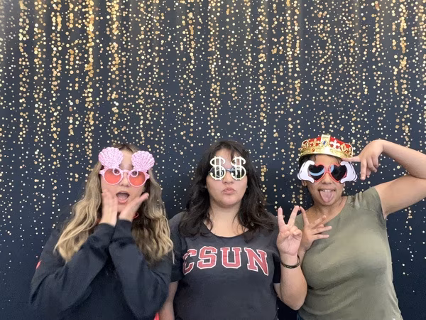 Three young women making silly faces in front of a black backdrop with gold bokeh sparkles at Cleveland HS. The young woman on the left wears pink seashell sunglasses with her mouth open in surprise and hands framing her cheeks. The young woman in the middle wears a grey CSUN t-shirt and silver dollar sign glasses, making a kissy face while throwing a peace sign. The young woman on the right wears an olive green shirt, a red and gold crown, and cartoon heart sunglasses, sticking her tongue out playfully while holding up peace signs.