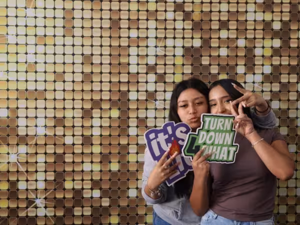 Two young women posing closely together in front of a sparkling gold sequin backdrop at Cleveland HS. The woman on the left is making a playful face while holding a purple "it's lit" sign, and the woman on the right is holding a green "TURN DOWN 4 WHAT" sign while throwing up a peace sign with her hand.