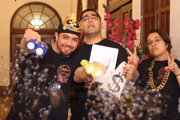 Three smiling event volunteers in matching black t-shirts shooting bubble guns toward the camera, posing playfully with a gold crown, a money bag, a thick gold chain, and a large Joker playing card.