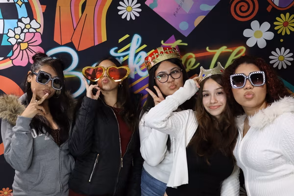 Five young women posing playfully with peace signs and kissy faces in front of a colorful 70s retro backdrop at Iglesia de Restauración Reseda. They are wearing fun props including studded square sunglasses, oversized red heart sunglasses, a large colorful crown, and a small gold tiara.