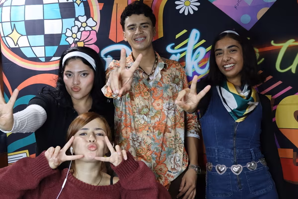 A group of four smiling teenagers posing with peace signs in front of a colorful 70s retro backdrop at Iglesia de Restauración Reseda. The group features a young man in a vintage floral shirt, a girl in a denim jumper with a heart belt, a girl with a white headband, and a girl in the front crouching and making double peace signs with a playful expression.